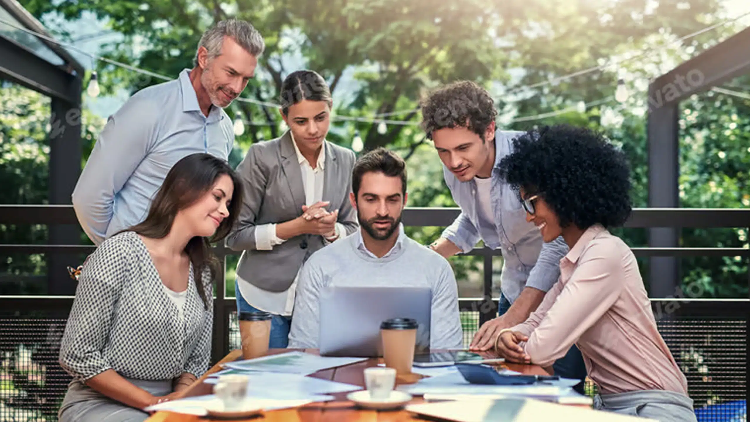 A diverse group of people huddled around a man on a laptop at a picnic table.